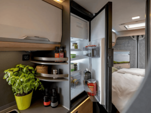 KNAUS BOXLIFE PLATINUM SELECTION 2027 – Kühlschrank im Camper Van Geöffneter Kühlschrank im KNAUS BOXLIFE PLATINUM SELECTION 2027 neben der Küchenzeile mit Blick zum Heckbett